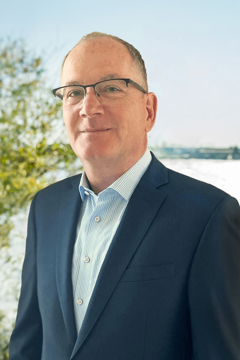 Mario Liebetrau, lächelnder Mann mit Brille in dunkelblauem Anzug und gestreiftem Hemd, steht vor einer neutralen Wand.